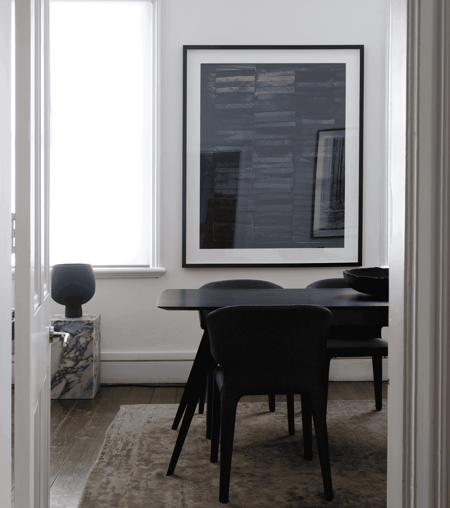 A meeting or dining room. Muted colors and light leaking in from a window. A black dining table sits in contrast, with a designed city-like painting in the background