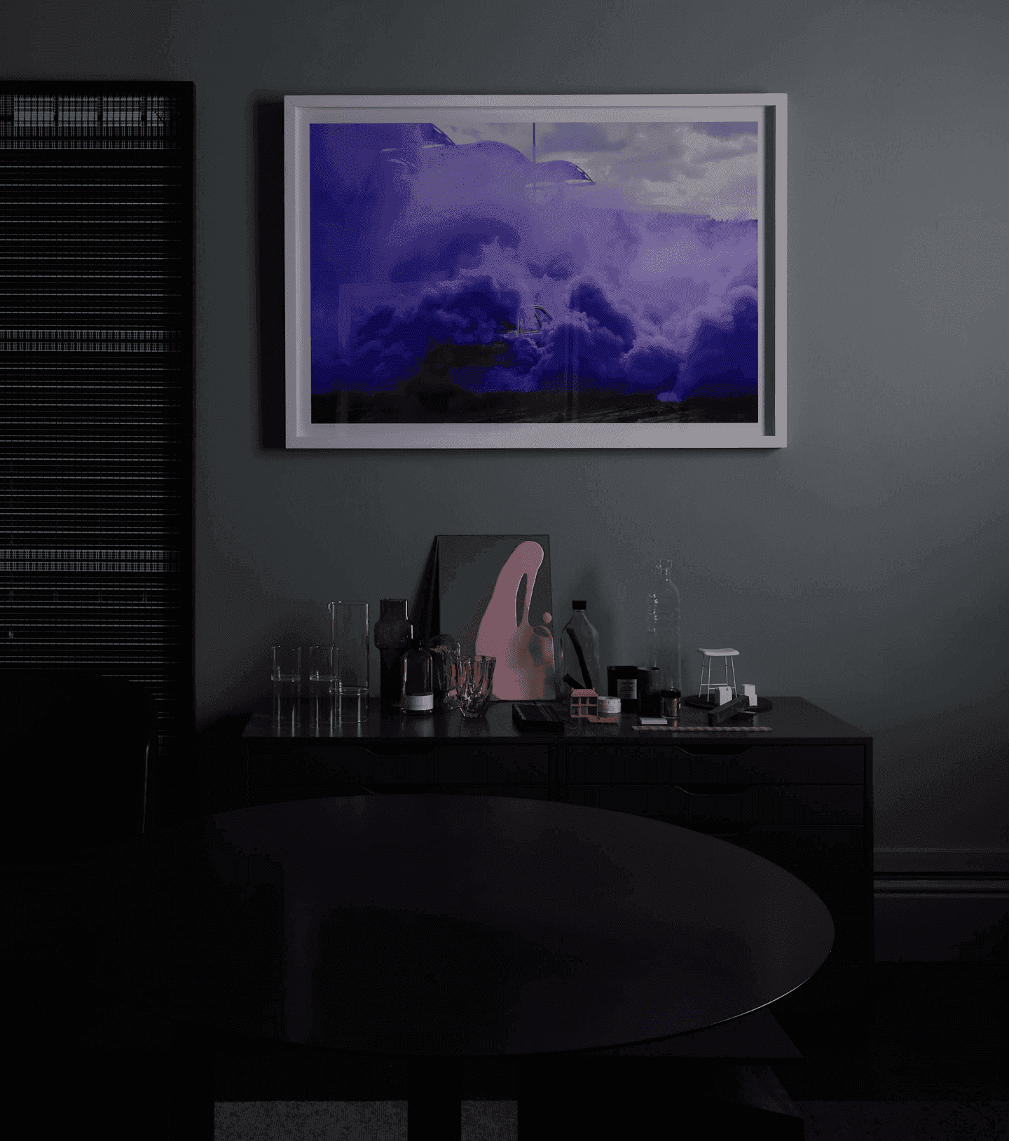 A dark moody picture of a side-table in black, on a dark grey wall. Small objects decorate it. The color comes from a purple-toned photo of a mountain